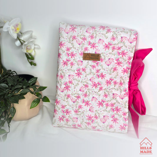 Floral-patterned book sleeve with a pink bow on a white surface, next to a plant and flowers.